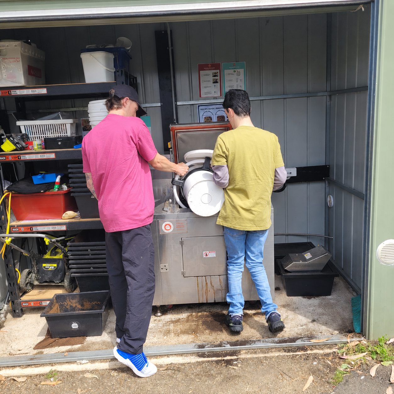 Two men pour a bucket into a large industrial dehydrator in a small shed